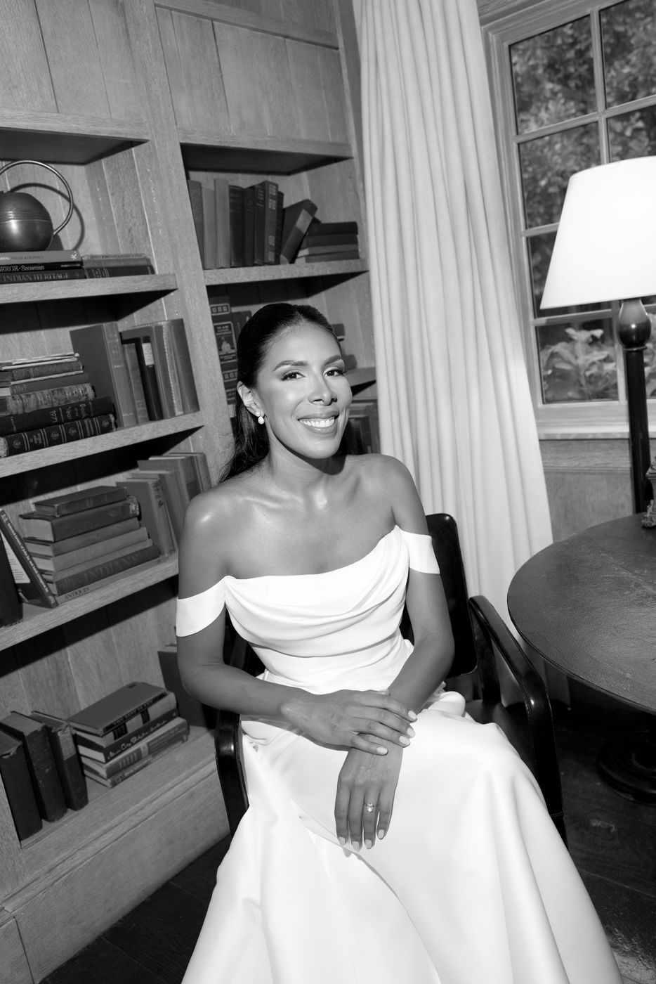 Black-and-white portrait of a smiling bride seated in a library, makeup by Doorbell Salon